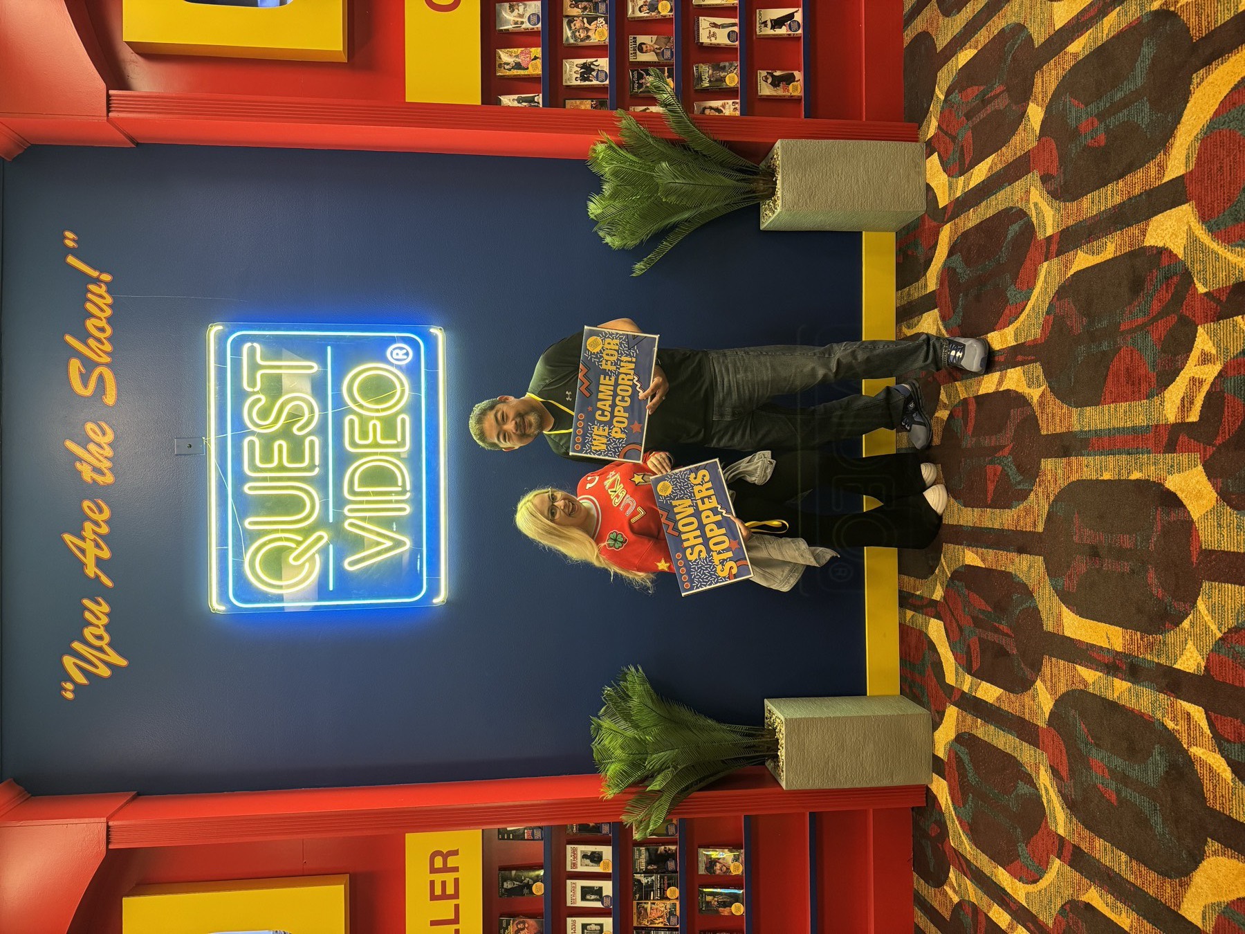 Lauryn and Richard Wang at Quest Video holding Show Stoppers and We Came For Popcorn signs under a neon sign
